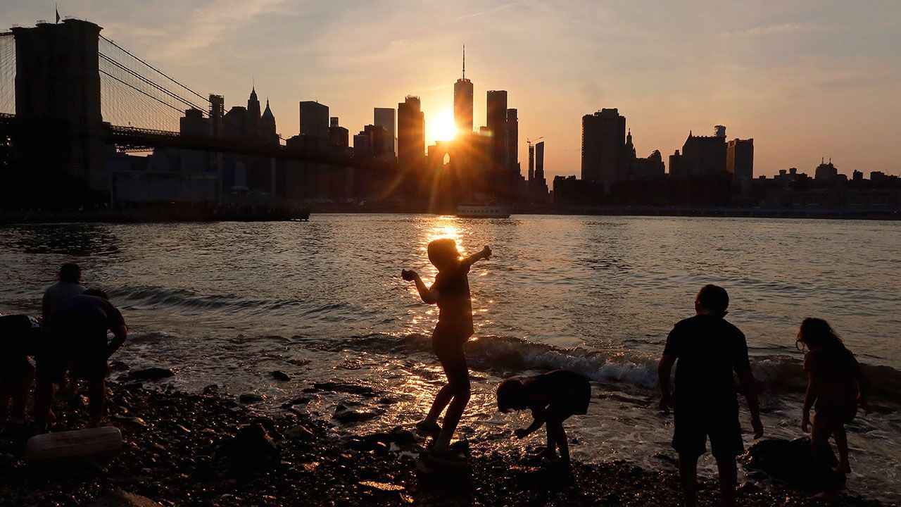A child throws rocks into the East River at Brooklyn Bridge Park as the sun sets behind the NYC skyline.