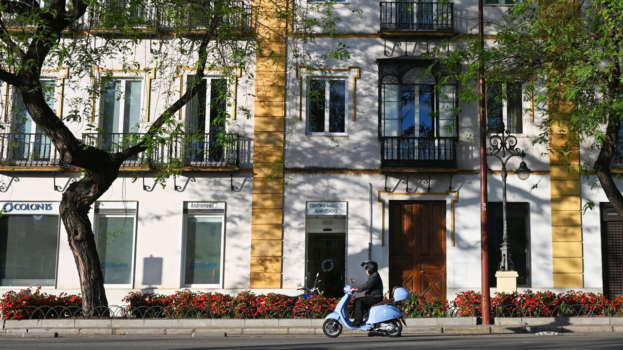 A man riding a Vespa scooter past residential buildings in Spain.