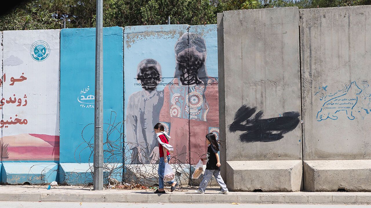 Two girls walk along Darul Aman Road in Kabul past a mural in which the human faces have been obscured with black paint in Kabul, Afghanistan, 2026.