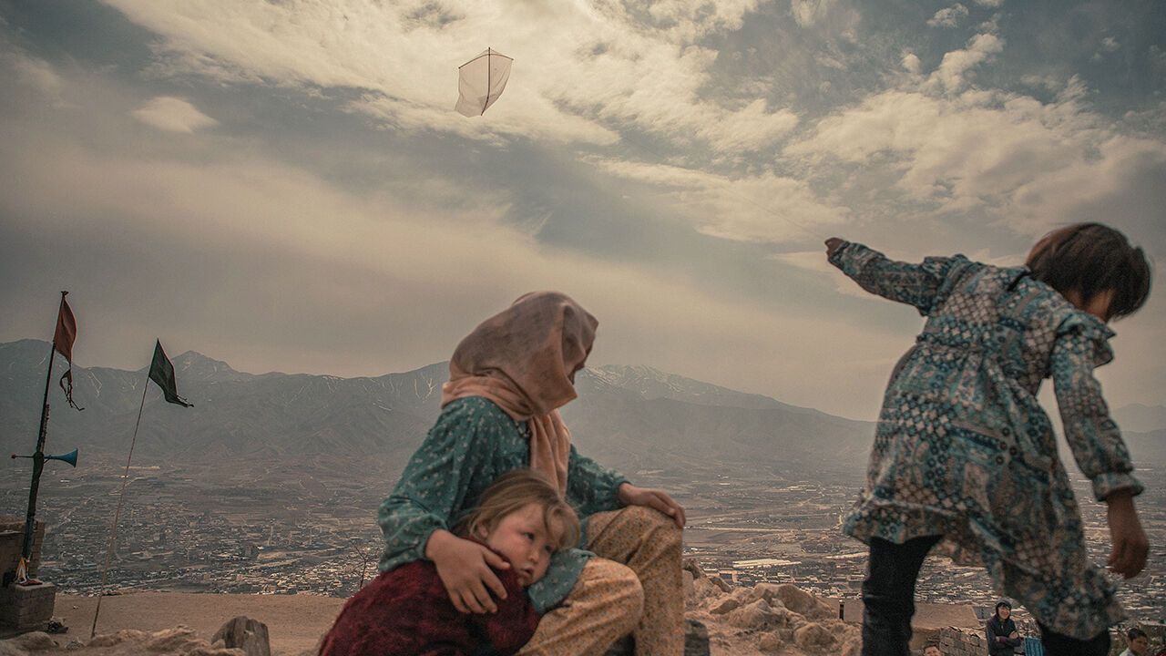 A woman sits with children on a hillside in Dasht-e-Barchi, while a girl watches a kite flying above western Kabul in 2026.