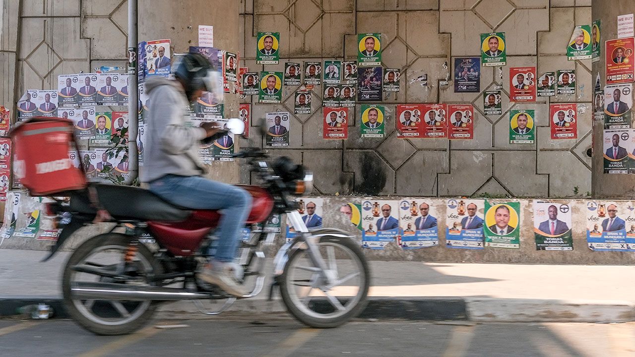 A motor bike delivery rider waits next a wall covered with electoral campaign posters in Lusaka, Zambia