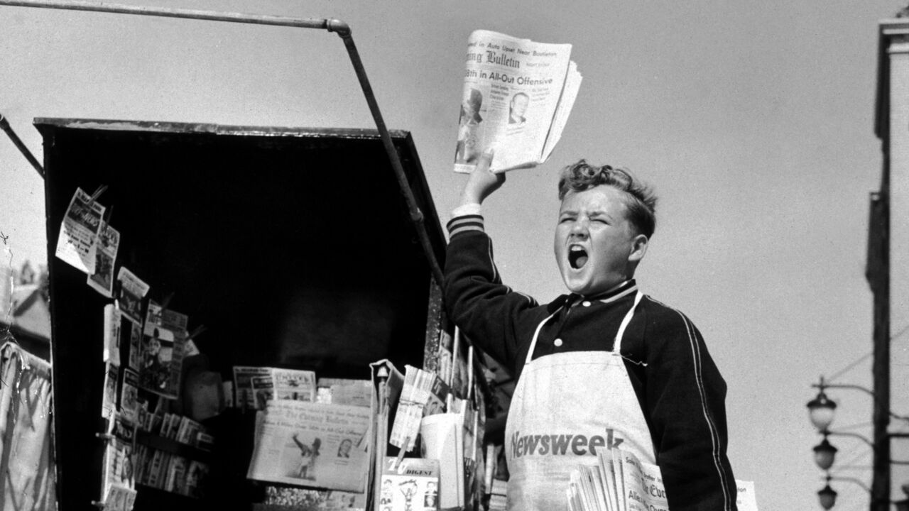 A newsboy shouting beside a newsstand while holding a newspaper in the air.