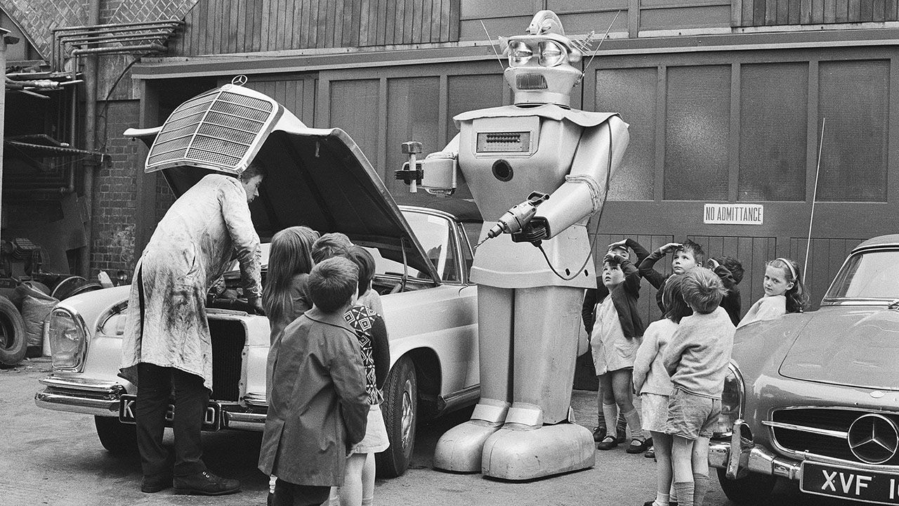 A nine foot high robot assists a garage mechanic while children from a nearby school look amazed.