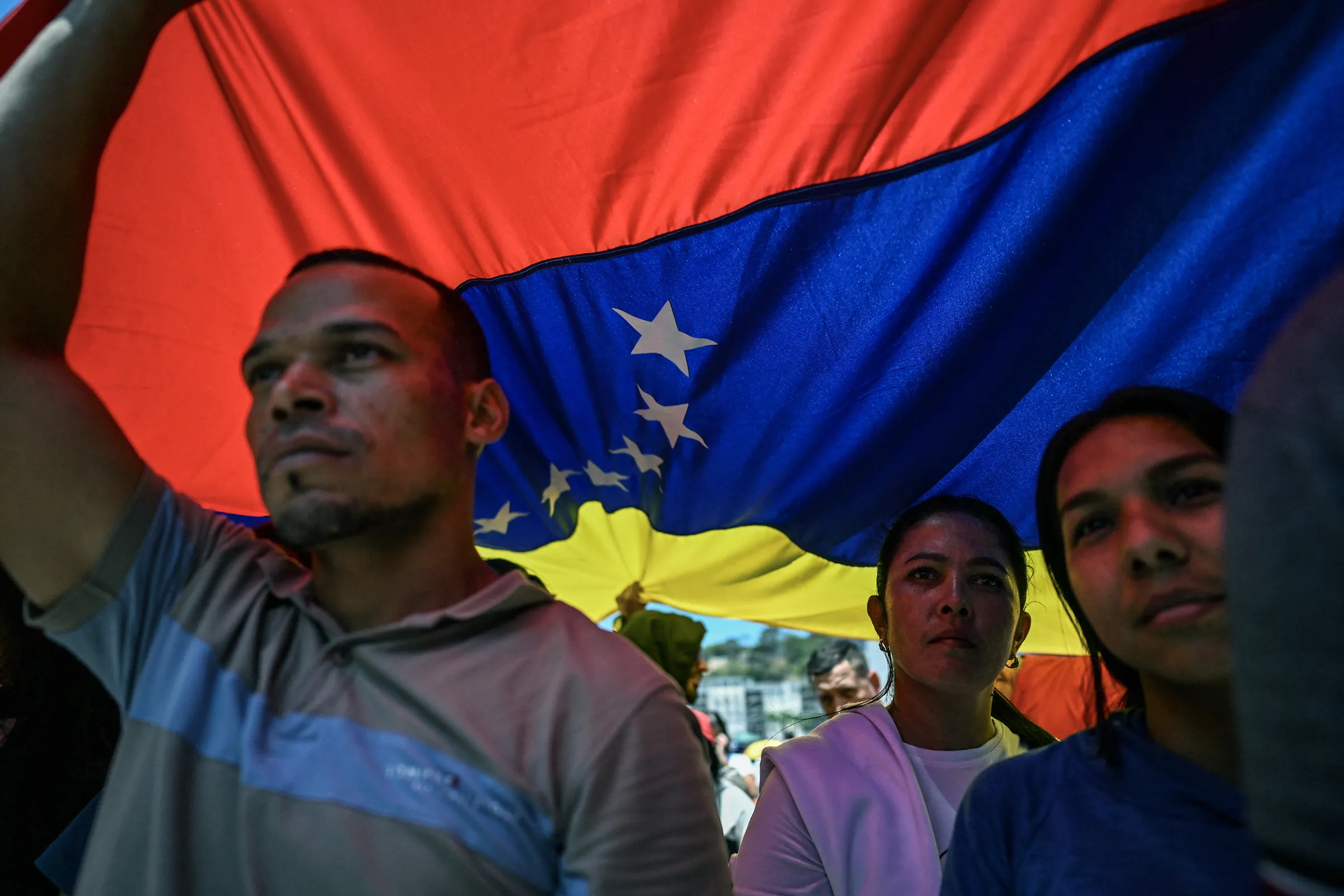 People walk under a huge Venezuelan flag during a march demanding the complete lifting of US sanctions in Caracas on March 23, 2026. (Photo by Juan BARRETO / AFP via Getty Images)